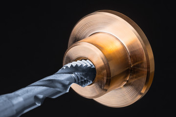 Detail of spiral flute tap in golden metal part. Artistic close-up of a silver-blue steel cutting tool for threading and bronze workpiece. Tapping on black background. Engineering industry, metalwork.