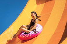 Young Pretty Woman On The Inflatable Ring Having Fun On The Orange Water Slide In The Aqua Park. Summer Vacation. Weekend On Resort