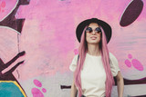 Fototapeta  - Happy stylish young hipster woman with long pink hair, hat and sunglasses on the street.