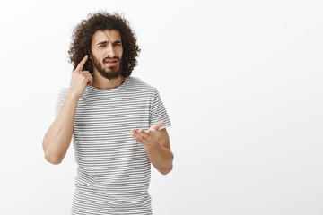 Wall Mural - What I cannot hear you, what do you want. Portrait of confused bothered handsome Eastern guy with afro hairstyle, directing at ear and pointing with palm, being in loud room over gray background