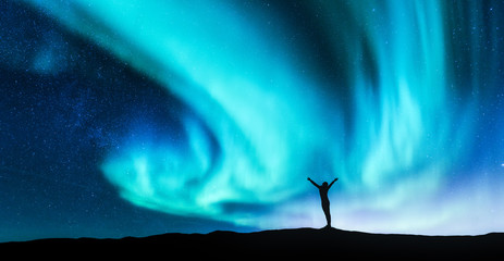 Wall Mural - Northern lights and silhouette of standing woman with raised up arms on the hill in Norway. Aurora borealis and happy girl. Starry sky, green polar lights. Night landscape. Travel background. Concept
