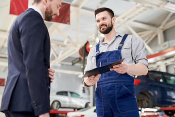 Low angle portrait of smiling bearded mechanic talking to businessman standing in clean car service and repair center