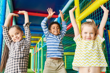 Wall Mural - waist up portrait of three happy kids jumping raising hands while having fun in play area and lookin