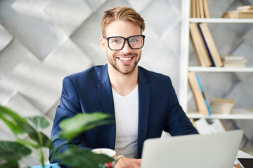 Wall Mural - Love my job. Waist up portrait of pleased man sitting at laptop with wide smile. He is looking forward wearing eyeglasses with delight and confidence