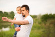 Dad keeps his son in arms and points with his hand towards something. Father and son together outdoors, dad shows his boy something ahead. Green background. Active family concept. Room for copy text