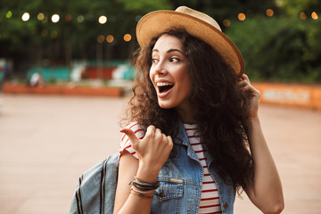 Wall Mural - Photo of excited pretty woman 18-20 with curly brown hair, laughing and pointing finger aside while walking in hipster spot or modern park with colorful lamps background