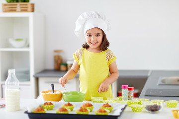 Wall Mural - family, cooking, baking and people concept - happy little girl in chefs toque making batter for muffins or cupcakes at home kitchen