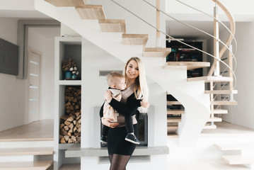 Wall Mural - Stylish young mom keeps in ergo backpack of her cute year-old baby at home in a beautiful interior Convenience for mom.