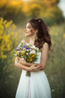 Beautiful lovely bride is wearing white fashion dress holding bouquet of wildflowers in yellow field, nature wedding concept