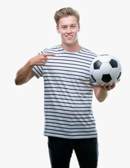 Sticker - Young handsome blond man holding soccer ball very happy pointing with hand and finger