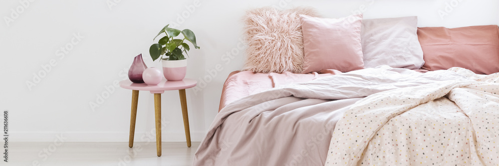 Real photo of a small table with a plant standing next to a bed with pink bedding in bedroom interior with white walls - obrazy, fototapety, plakaty 