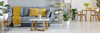 Real photo of a grey sofa with yellow pillows standing behind a table and next to a shelf with ornaments in living room interior with table and chairs in the bacground