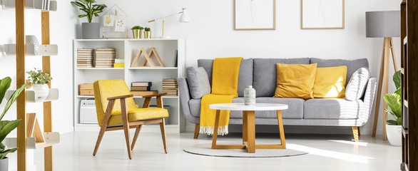 Real photo of a couch with yellow pillows standing bext to shelves with books and ornaments and lamp, and behind a table and armchair in spacious living room interior with posters on a wall