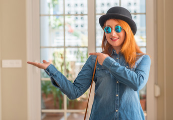 Sticker - Stylish redhead woman wearing bowler hat and sunglasses amazed and smiling to the camera while presenting with hand and pointing with finger.