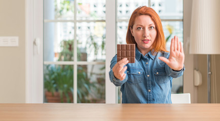 Wall Mural - Redhead woman holding chocolate bar at home with open hand doing stop sign with serious and confident expression, defense gesture