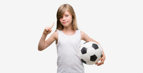 Sticker - Young blonde toddler holding football ball surprised with an idea or question pointing finger with happy face, number one