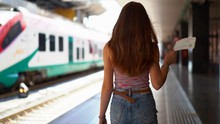 Woman Traveler At The Station Goes To The Platform On The Background Of A High-speed Train Going Along The Road. The Tourist Keeps The Ticket On The Train In His Hand. Train Station Concept