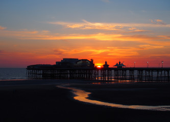 scenic view of blackpool north pier in glowing red evening light at sunset with illuminated pink and yellow sky and clouds with colours reflected on the dark beach in structure in silhouette