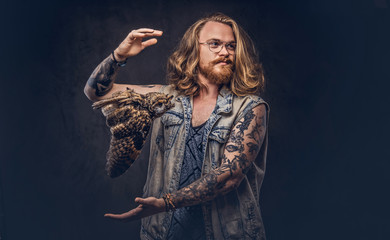 Wall Mural - Portrait of a tattoed redhead hipster male with long luxuriant hair and full beard dressed in a t-shirt and jacket holds a keeps the scarecrow of an owl in a studio. Isolated on the dark background.