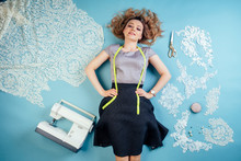 Attractive Woman Seamstress Tailor (dressmaker) Dreams And Thinks About A New Collection Of Clothes On The Floor With Sewing Machine And Measuring Tape On A Blue Background In The Studio Top View .