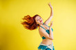 beautiful red-haired woman dancing oriental belly dance in blue suit on yellow background in studio