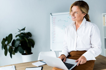 Wall Mural - attractive businesswoman sitting on table with laptop and looking away in office