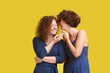 © Rithor - Cheerful curly hairstyle girls gossip. Charming caucasian woman tells funny story to her best friend in ear, joke causes a smile and laughter. Communication concept, friendship, yellow background