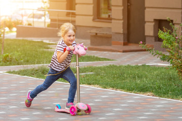 cute little girl riding scooter in city park on bright summer day. blond toddler having fun on stree