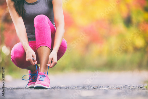 Runner tying running shoes laces in autumn background. Athlete woman getting ready to run race in fall landscape with yellow leaves foliage in park.