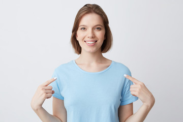 Poster - Happy confident young woman in blue t shirt smiling and pointing at herself with fingers on both hands isolated over white background