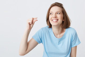 Poster - Cheerful beautiful young woman in blue t shirt laughing and showing tiny size by fingers of her hand isolated over white background