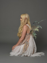 Full Length Portrait A Blonde Girl Wearing Fairy Costume. Seated Pose, On Grey Studio Background.