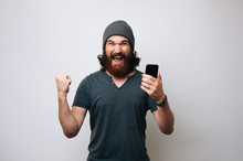 Cheerful Young Bearded Man Wearing Grey T-shirt And Fur Cap Hat Is Surprised And Happy. Winner Man With Smartphone Showing Winner Gesture On White Background.