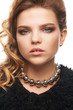 Close up portrait of a young fair-haired lady with one sided hairstyle. The girl in a black shaggy coat and a beaded collar necklace, posing against the white background, looking at the camera.