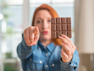 Wall Mural - Redhead woman holding chocolate bar at home pointing with finger to the camera and to you, hand sign, positive and confident gesture from the front