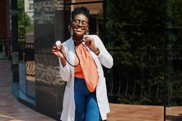 Wall Mural - African american doctor female at lab coat, glasses with stethoscope posed outdoor against clinic.