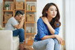 © DragonImages - Sad Asian woman sitting alone with man on background being in conflict at home