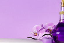 Pale Purple Orchid On A White Table With Purple Background. On The Table Is A Fragrant Candle And A Purple Bottle