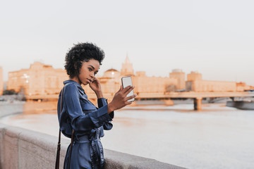 cute young african-american female tourist in a blue dress is photographing herself near the city ri