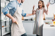 excited girlfriend standing with hands up and boyfriend holding banana gun in kitchen