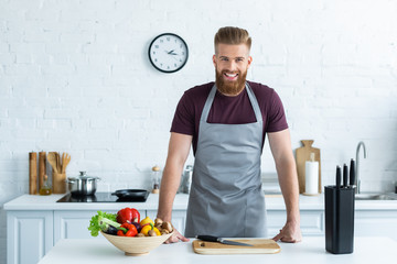 Wall Mural - handsome bearded young man in apron smiling at camera while cooking in kitchen