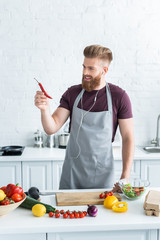 Wall Mural - handsome smiling bearded man in apron listening music in earphones and holding chili pepper while cooking in kitchen