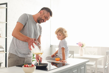 smiling man pouring dough into frying pan while his son sitting near on tabletop at kitchen