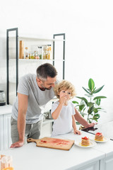 Wall Mural - smiling father looking how his son eating strawberry near tabletop with pancakes at kitchen