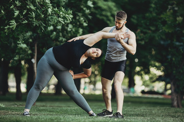 Personal trainer correcting his client at street workout. Overweight woman doing exercise with instructor support.