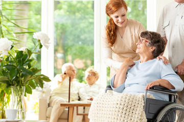 Wall Mural - Tender caregiver saying goodbye to an elderly pensioner in a wheelchair in a day care facility.  A companion pushing the wheelchair. Other elderly people in the blurred background.