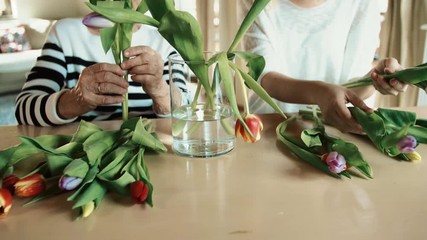 Wall Mural - Elderly grandmother with an adult granddaughter at home., putting flowers in a vase.