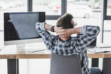 back view of programmer sitting with hands behind head at workplace