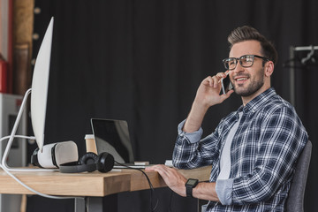 smiling young man in eyeglasses talking by smartphone and looking away at workplace