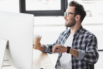 handsome smiling young programmer in eyeglasses holding paper cup while working with desktop computer